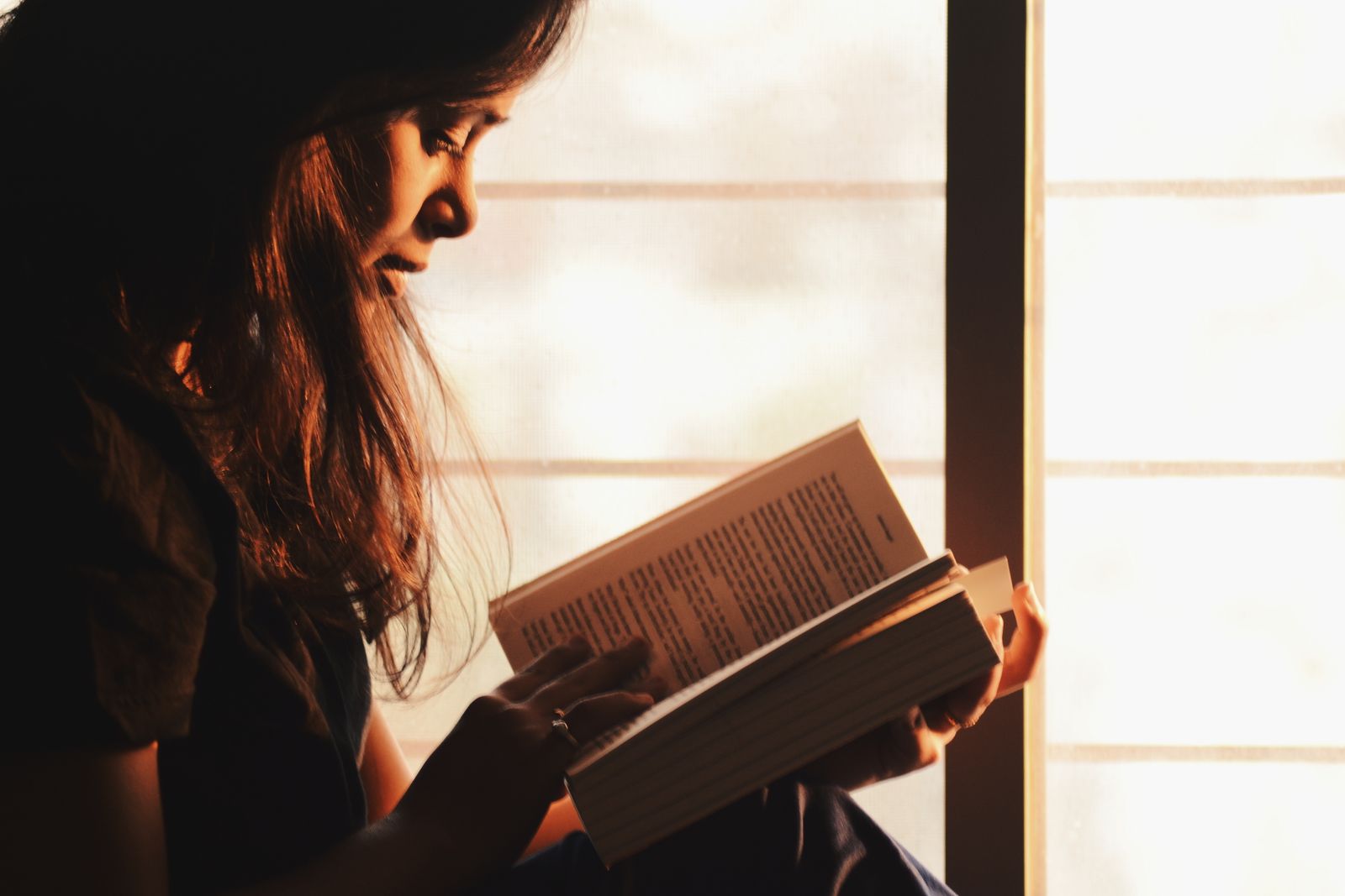 A person reading a book with calm, absorbed focus by a window