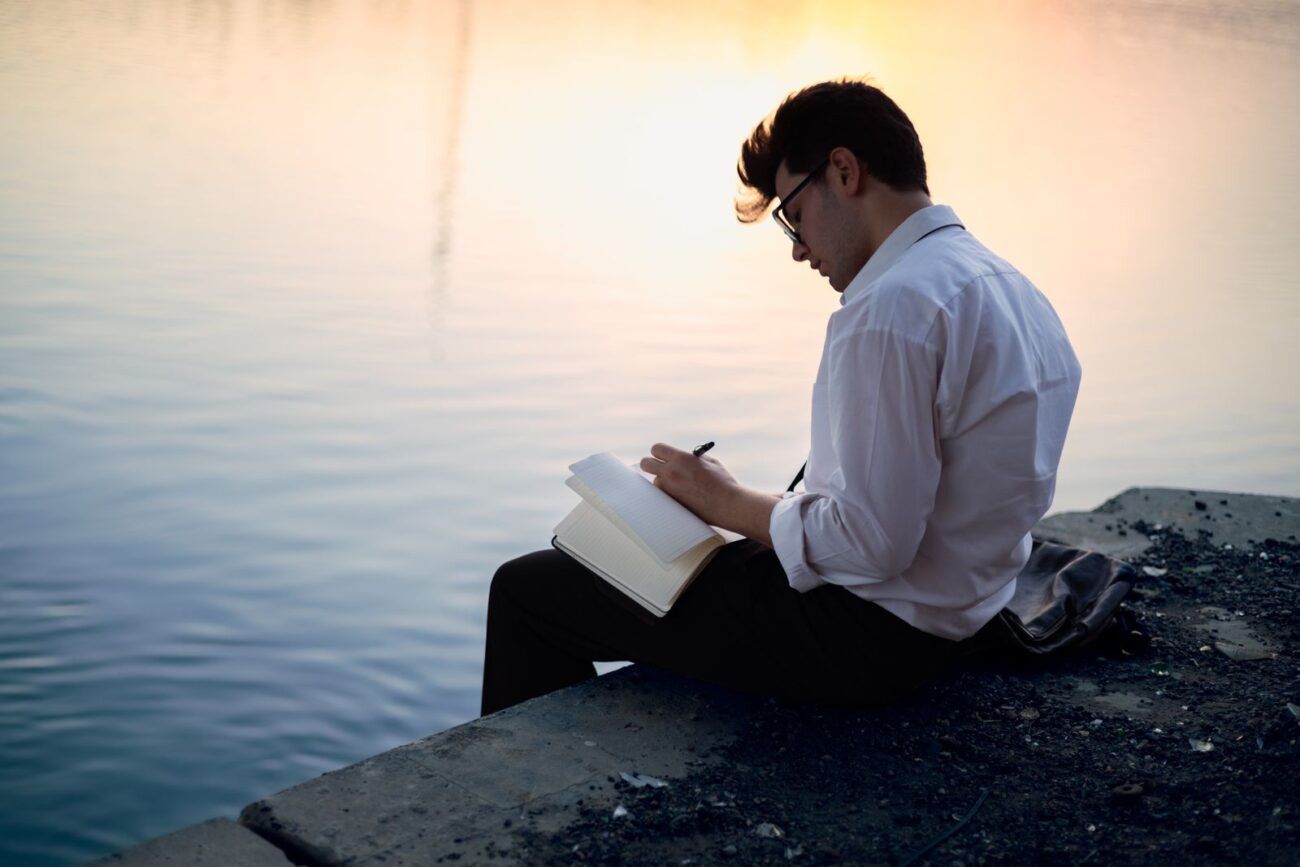A person sitting by the water at sunset, writing in a notebook