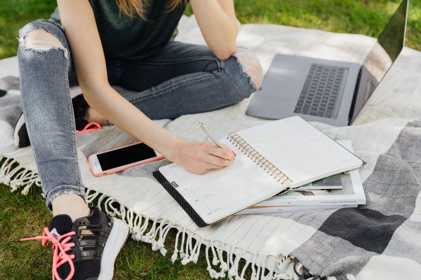 A person writing in a notebook outdoors on a blanket with a laptop