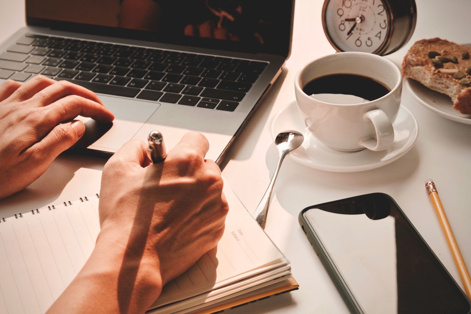 Hands writing in a notebook beside a laptop, coffee, a clock and a phone