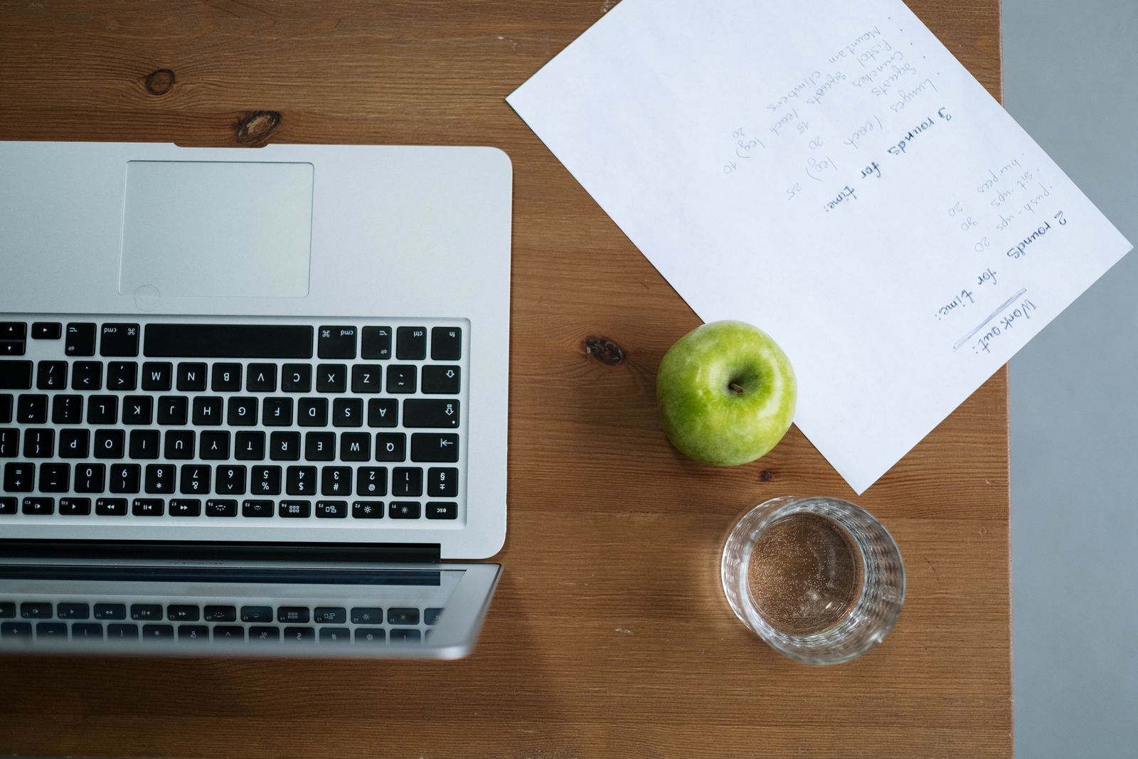 Top-down view of a wooden desk featuring a laptop, paper notes, a green apple, and a glass of water, symbolizing structured daily habits and routines.