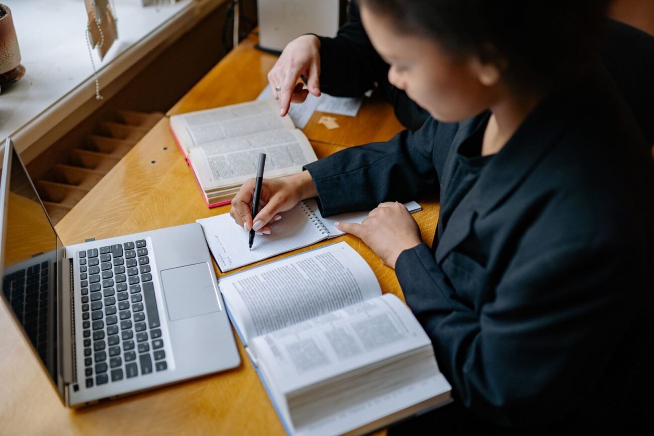 A person taking notes while studying with an open book and laptop